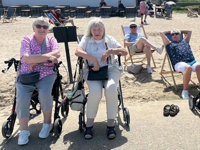 Zwei Senioren sitzen mit Rollatoren am Strand bei sonnigem Wetter.
